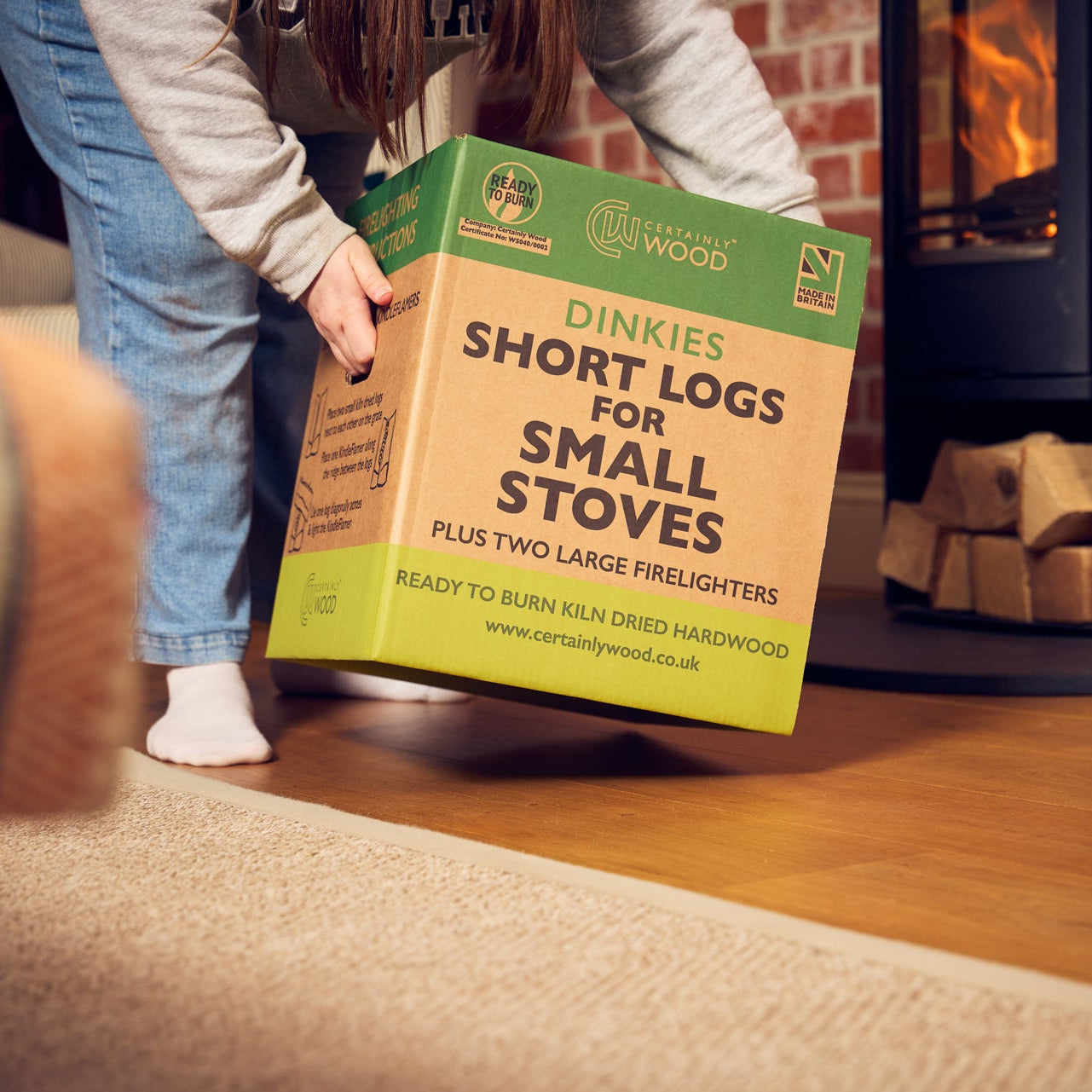Person holding a box of Dinkies Short Logs for Small Stoves in front of a lit fireplace.